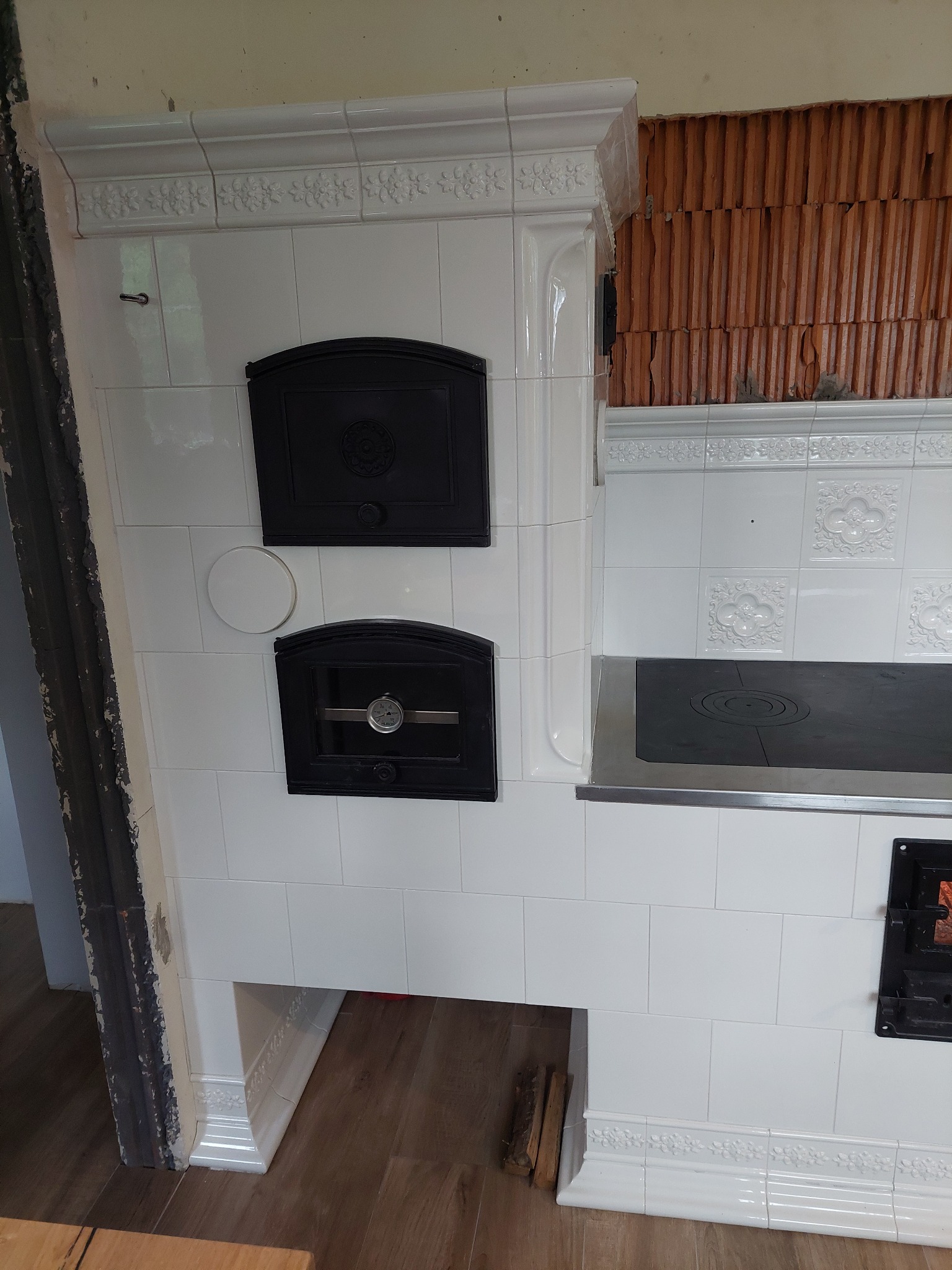 Biały piec kaflowy z czarnymi drzwiczkami i płytą grzewczą, widoczne cegły w tle. Dekoracyjne zdobienia na kaflach i listwach. Częściowo widoczne drewno na podłodze.