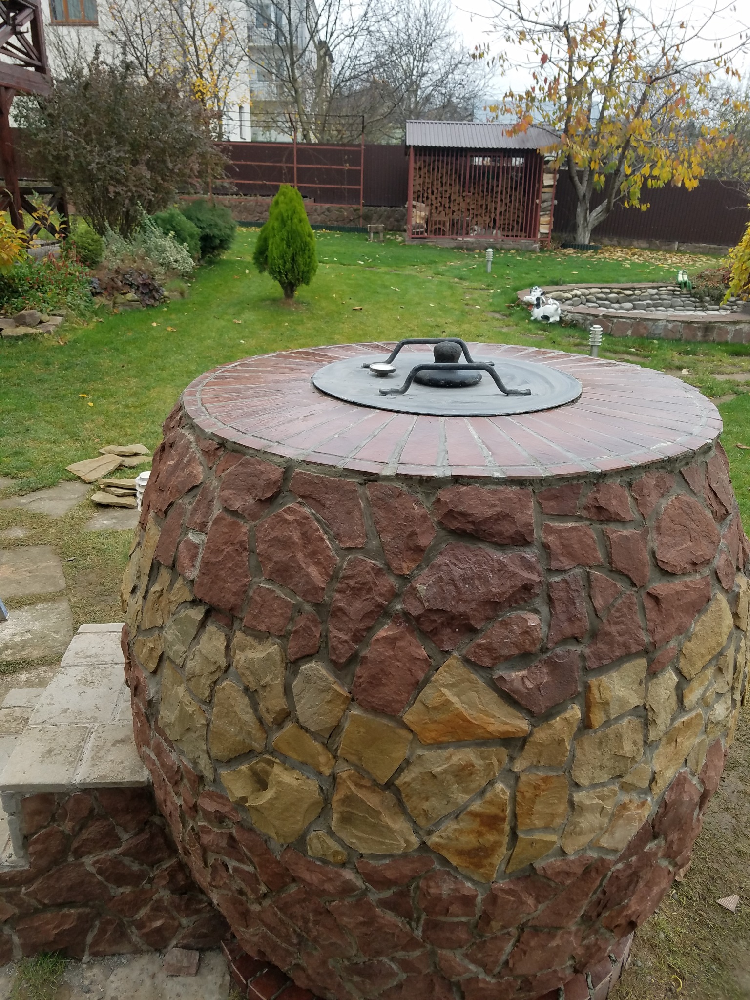 Okrągły piec ogrodowy z kamienia i cegły, z metalową pokrywą na trawniku. W tle widoczny ogród z drzewami i budynkiem.