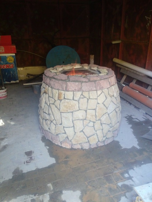 Ognisko ogrodowe z kamienia w rustykalnym stylu, z widocznym płomieniem wewnątrz. Tło stanowią materiały budowlane i ściana w ciemnych odcieniach.