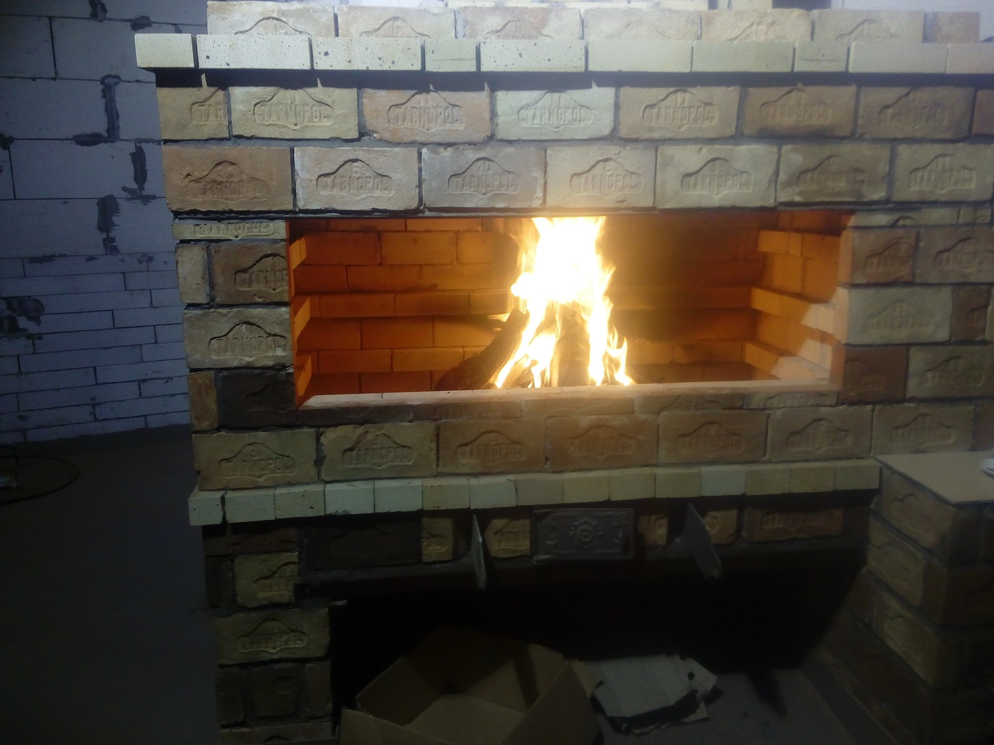 Murowany kominek z cegieł z paleniskiem, w którym płonie ogień. Cegły z widocznym napisem 'CZARNOPOL'. Widoczne niedoskonałości i surowy stan wykończenia.