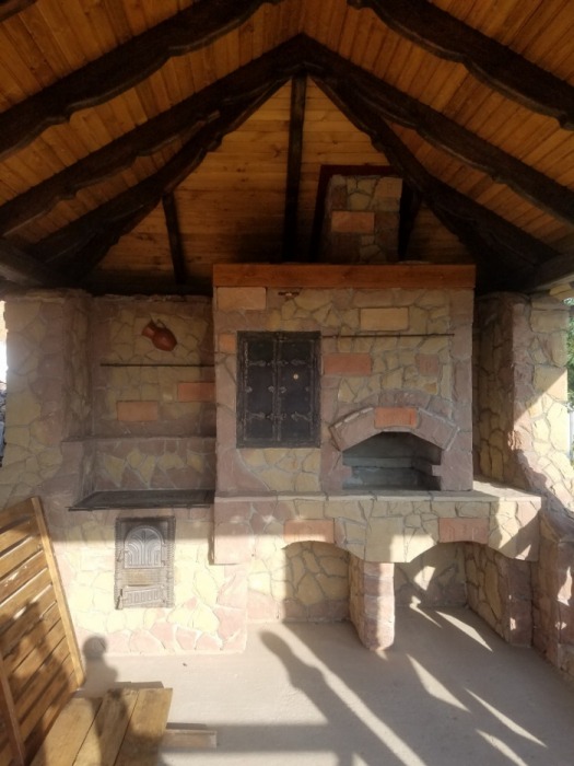 Kamienny piec chlebowy z paleniskiem i wędzarnią pod drewnianą wiatą. Rustykalny styl, widoczne detale wykończenia i oświetlenie słoneczne.