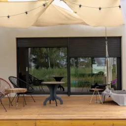 Zadaszony taras z drewnianą podłogą, wyposażony w meble ogrodowe, wiszące lampiony i zasłony, z widokiem na ogród przez okno z opuszczoną roletą zewnętrzną.