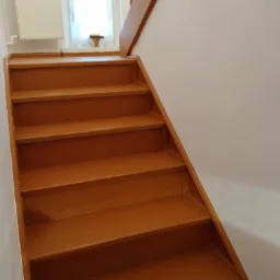 Odnowione drewniane schody w kolorze miodowym prowadzące do góry; biała ściana i okno z firanką w tle.