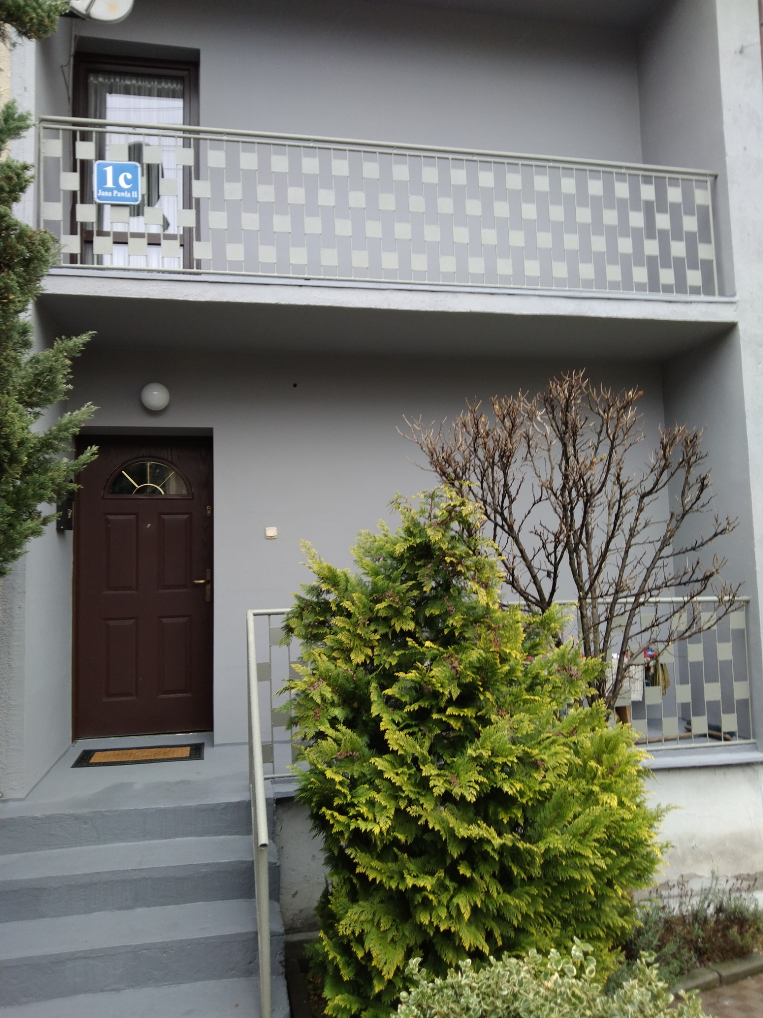 Szare wejście do budynku z brązowymi drzwiami, schodami i zielonymi krzewami. Nad drzwiami balkon z szarą balustradą i tabliczką z adresem.