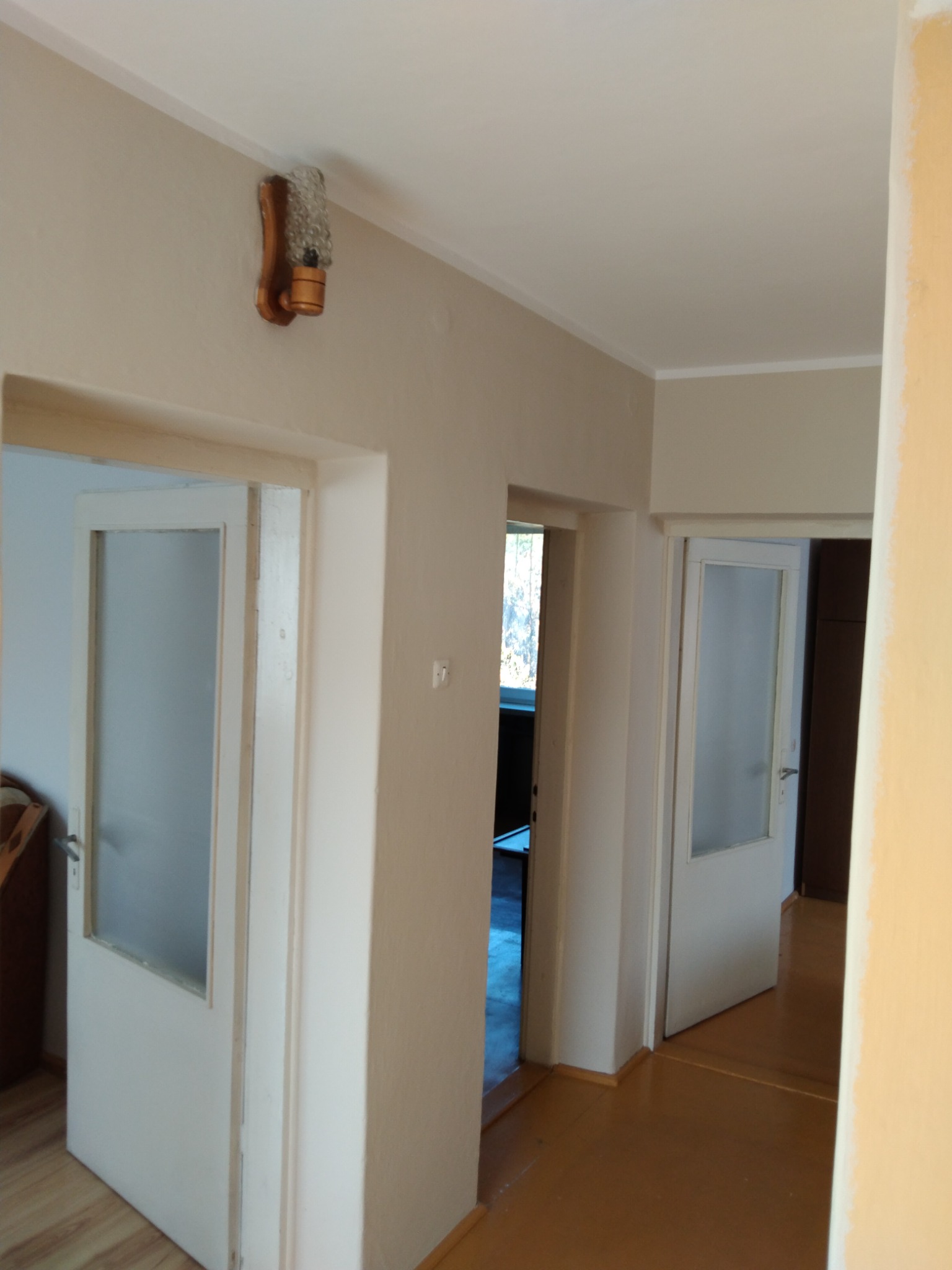 Korytarz po malowaniu z trzema parami drzwi, widoczne ślady świeżej farby na ścianie po prawej stronie i drewniany kinkiet z szklanym kloszem na ścianie