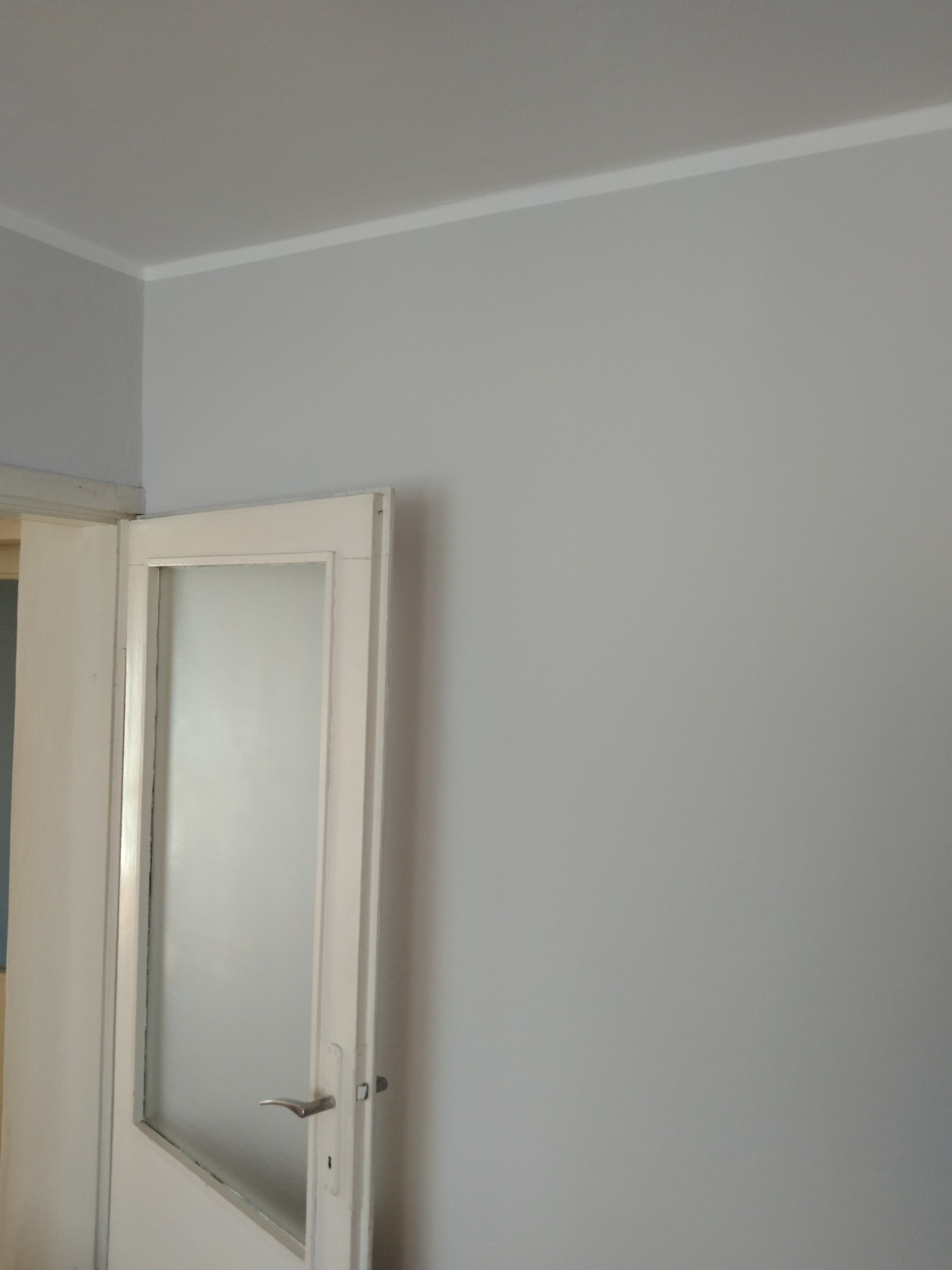 Świeżo pomalowana szara ściana w pokoju z białą listwą przysufitową i częściowo otwartymi, białymi drzwiami z matową szybą i srebrną klamką.