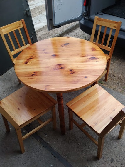 Okrągły stół z drewna sosnowego i cztery krzesła o prostej konstrukcji ustawione na tle otwartych drzwi samochodu dostawczego.