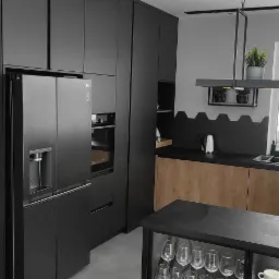 Nowoczesna kuchnia z czarnymi matowymi frontami, drewnianymi akcentami i heksagonalnymi płytkami na ścianie, oświetlona lampą wiszącą z doniczkami, z widocznym sprzętem AGD marki LG.