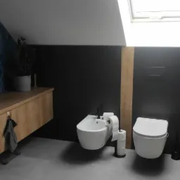 Nowoczesna łazienka na poddaszu z drewnianą szafką pod umywalką, bidetem i toaletą wiszącą na tle czarnej ściany z drewnianym akcentem i niebieskiej ściany z geometrycznym wzorem.