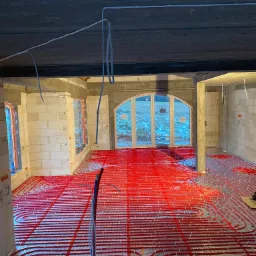 Wnętrze w stanie surowym z widoczną instalacją ogrzewania podłogowego z czerwonych rur ułożonych na folii, widoczne okna z widokiem na zasnieżony ogród.