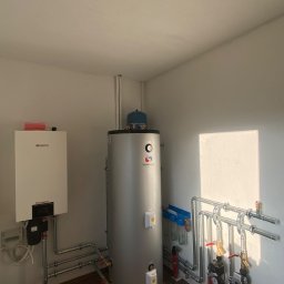 CHIMEX Instalacje Sanitarne - Zmontowany system grzewczy w kotłowni: biały piec gazowy, srebrny zasobnik ciepłej wody, rury i zawory. Widoczne filtry wody i osprzęt. Oświetlenie dzienne.