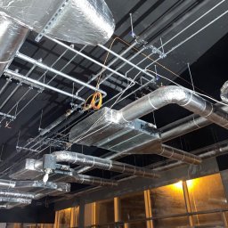 CHIMEX Instalacje Sanitarne - Skomplikowana sieć srebrnych rur wentylacyjnych i instalacji pod sufitem, widoczne elementy mocujące i izolacja, w tle okna z widokiem na zewnątrz.