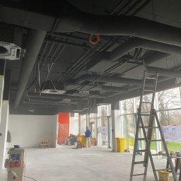 CHIMEX Instalacje Sanitarne - Wnętrze w trakcie prac instalacyjnych: widoczne rury pod sufitem, drabina, narzędzia i ekipa w oddali. Duże okna wpuszczają światło dzienne. Etap prac instalacyjnych.