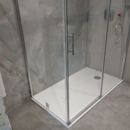 Robert Gembala - Nowoczesna kabina prysznicowa z białą brodzikiem i szklanymi ścianami na tle płytek imitujących marmur.