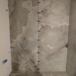 Robert Gembala - Montaż wielkoformatowych płytek imitujących marmur na ścianie łazienki, z widocznymi krzyżykami dystansowymi i narzędziami na podłodze.