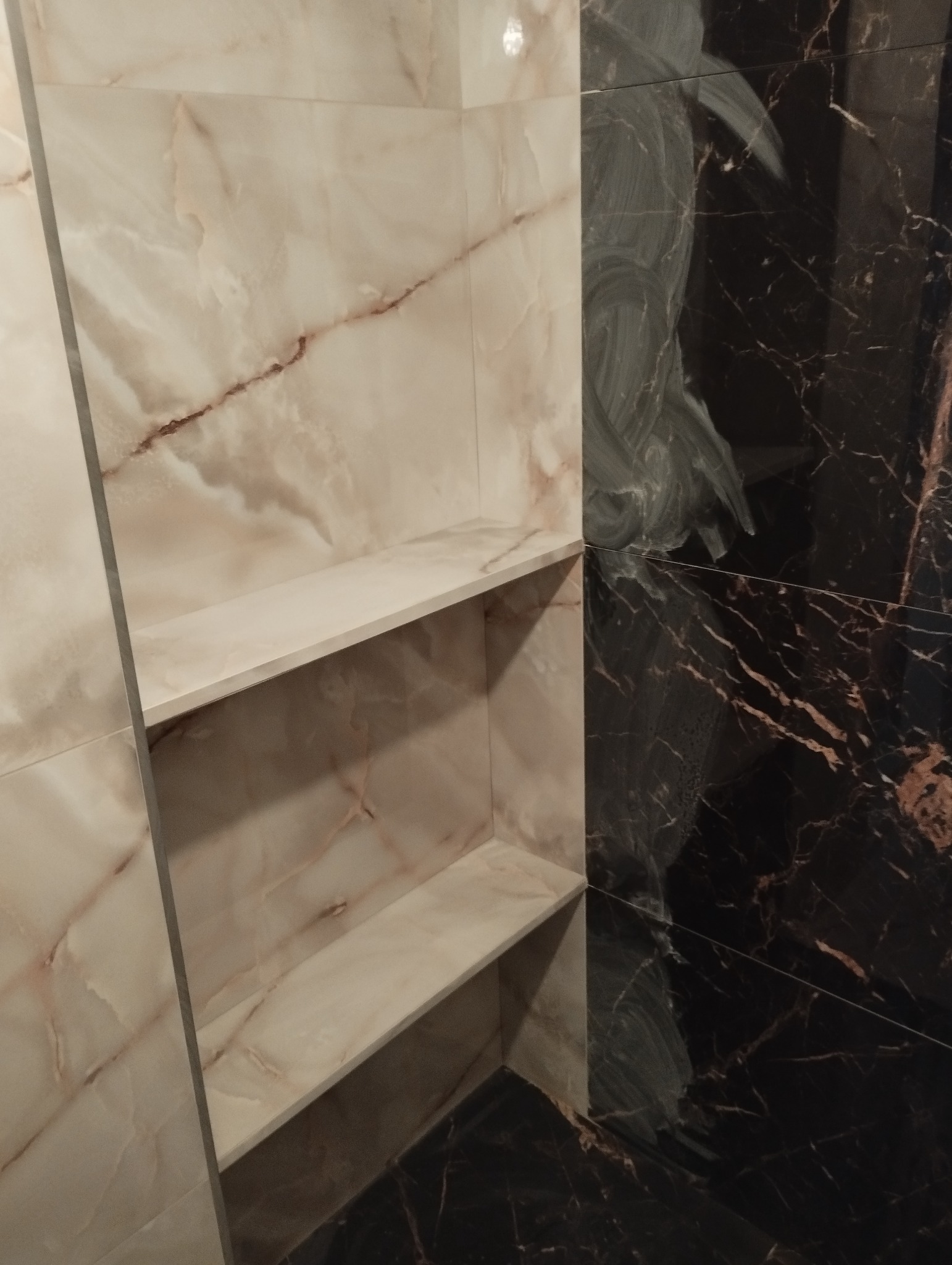 Wnęka z półkami wyłożona płytkami imitującymi marmur w odcieniach beżu i brązu, sąsiadująca z czarną ścianą również wykończoną płytkami marmuropodobnymi.
