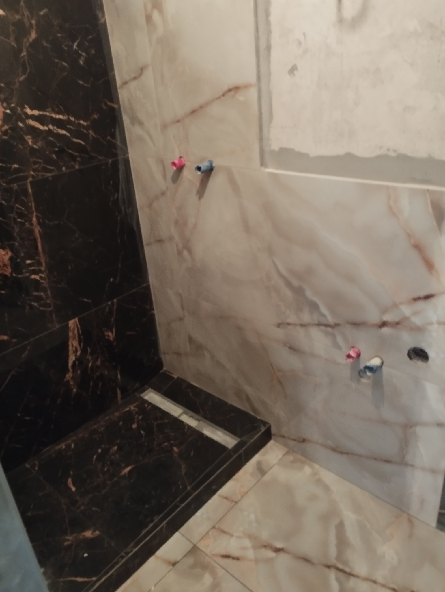 Fragment łazienki w trakcie wykańczania, widoczne płytki imitujące marmur w odcieniach bieli i brązu, czarny brodzik z odpływem liniowym, oraz wystające rurki instalacji wodnej i otwór pod baterię.