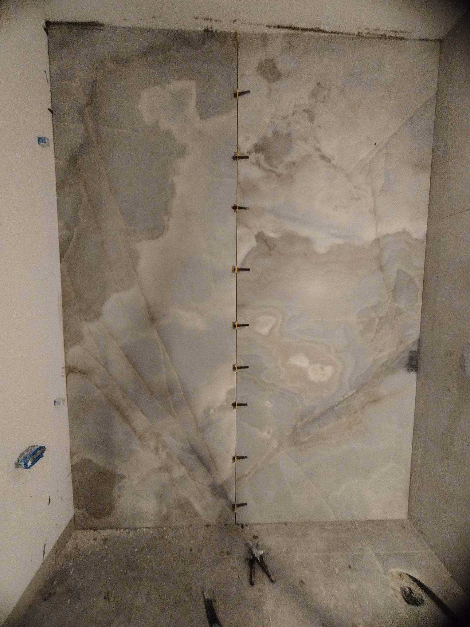 Montaż wielkoformatowych płytek imitujących marmur na ścianie łazienki, z widocznymi krzyżykami dystansowymi i narzędziami na podłodze.