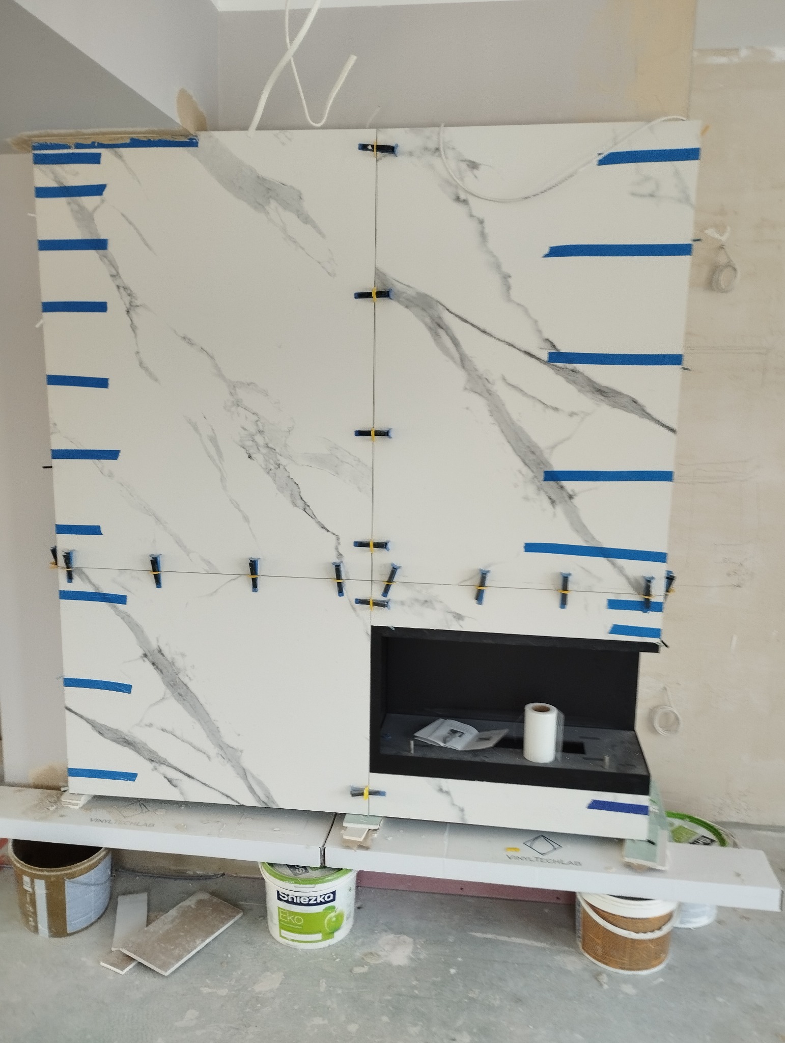 Montaż płytek imitujących marmur na ścianie z wnęką, zabezpieczonych niebieską taśmą i krzyżykami dystansowymi, z widocznymi pojemnikami z farbą i płytkami na podłodze.
