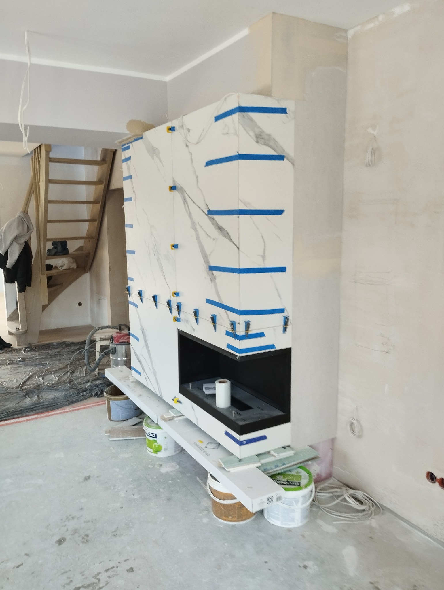 Montaż obudowy kominka z płytek imitujących marmur, zabezpieczonych niebieską taśmą i poziomowanymi krzyżykami dystansowymi, w surowym wnętrzu z widocznymi schodami i przewodami.