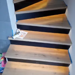 Schody drewniane z podświetleniem LED, zabezpieczone niebieską taśmą malarską, na jednym ze stopni leżą narzędzia i instrukcja montażu.