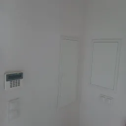 Biały panel alarmowy z podświetlonym wyświetlaczem, zamontowany na ścianie obok włącznika światła, z dwoma białymi, zabudowanymi skrzynkami rewizyjnymi w tle.