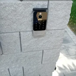 Nowoczesny wideodomofon z klawiaturą numeryczną zamontowany na szarym, kamiennym słupku ogrodzenia.