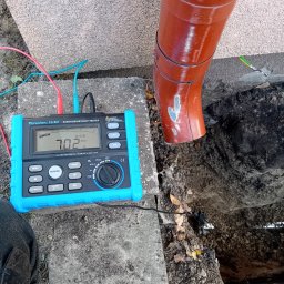 ELEKTRODAKO Usługi Elektryczne Daniel Dakowicz - Miernik rezystancji uziemienia Forscher FS 907 z podłączonymi przewodami pomiarowymi, wskazujący wartość 7.02 Ω, umieszczony na betonowej płycie obok rury spustowej z widocznym uziomem.