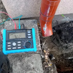 Miernik rezystancji uziemienia Forscher FS 907 z podłączonymi przewodami pomiarowymi, wskazujący wartość 7.02 Ω, umieszczony na betonowej płycie obok rury spustowej z widocznym uziomem.
