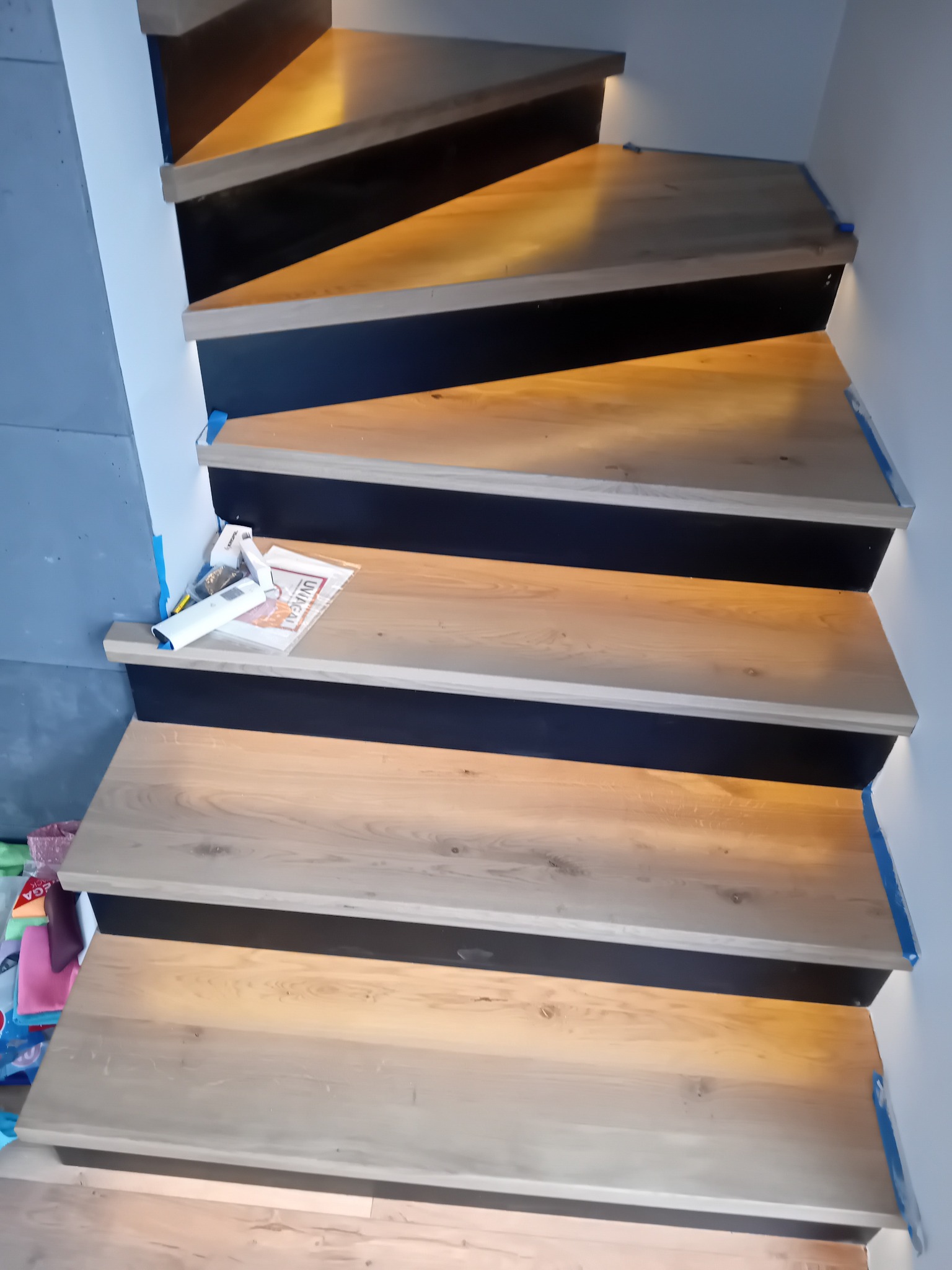 Schody drewniane z podświetleniem LED, zabezpieczone niebieską taśmą malarską, na jednym ze stopni leżą narzędzia i instrukcja montażu.