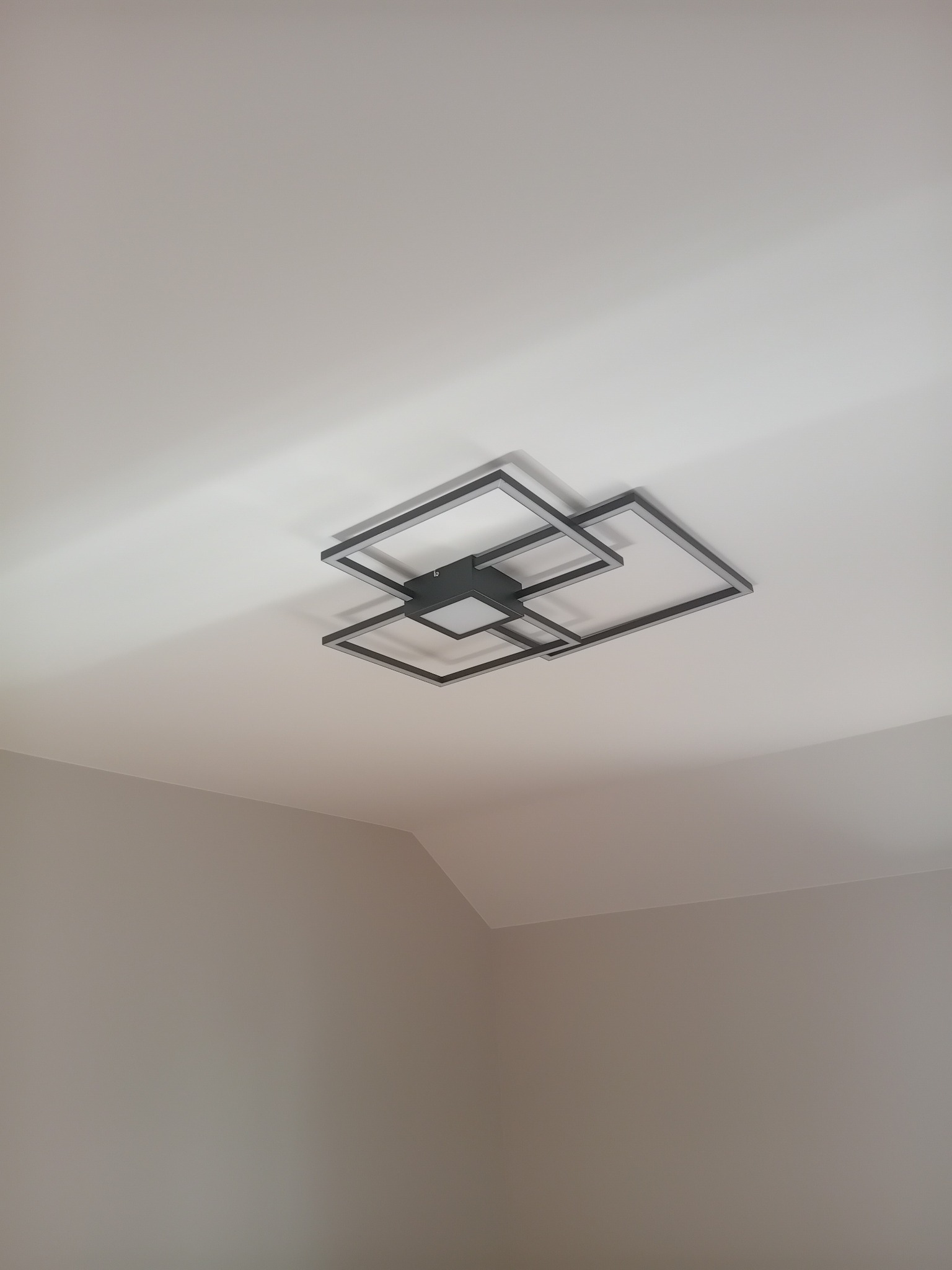 Czarna, nowoczesna lampa sufitowa LED o geometrycznym designie, składająca się z kwadratowych ram, zamontowana na białym suficie w pomieszczeniu o jasnych ścianach.