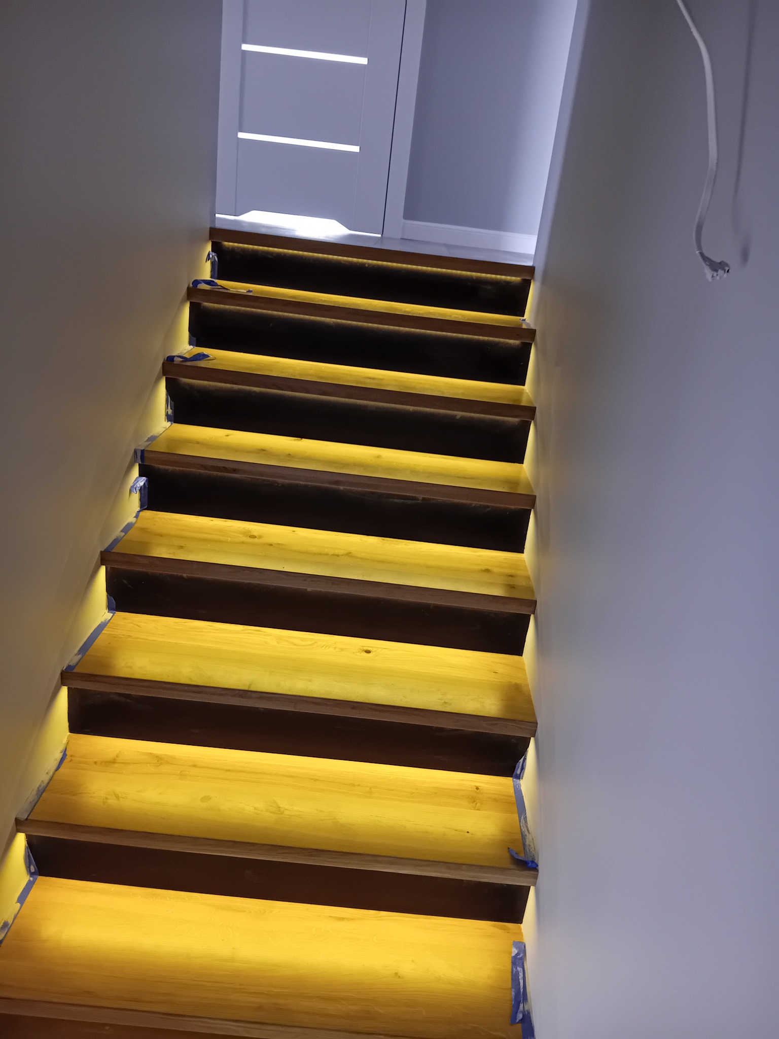 Podświetlone, drewniane schody w trakcie montażu oświetlenia LED, zabezpieczone niebieską taśmą malarską, widoczne kable.