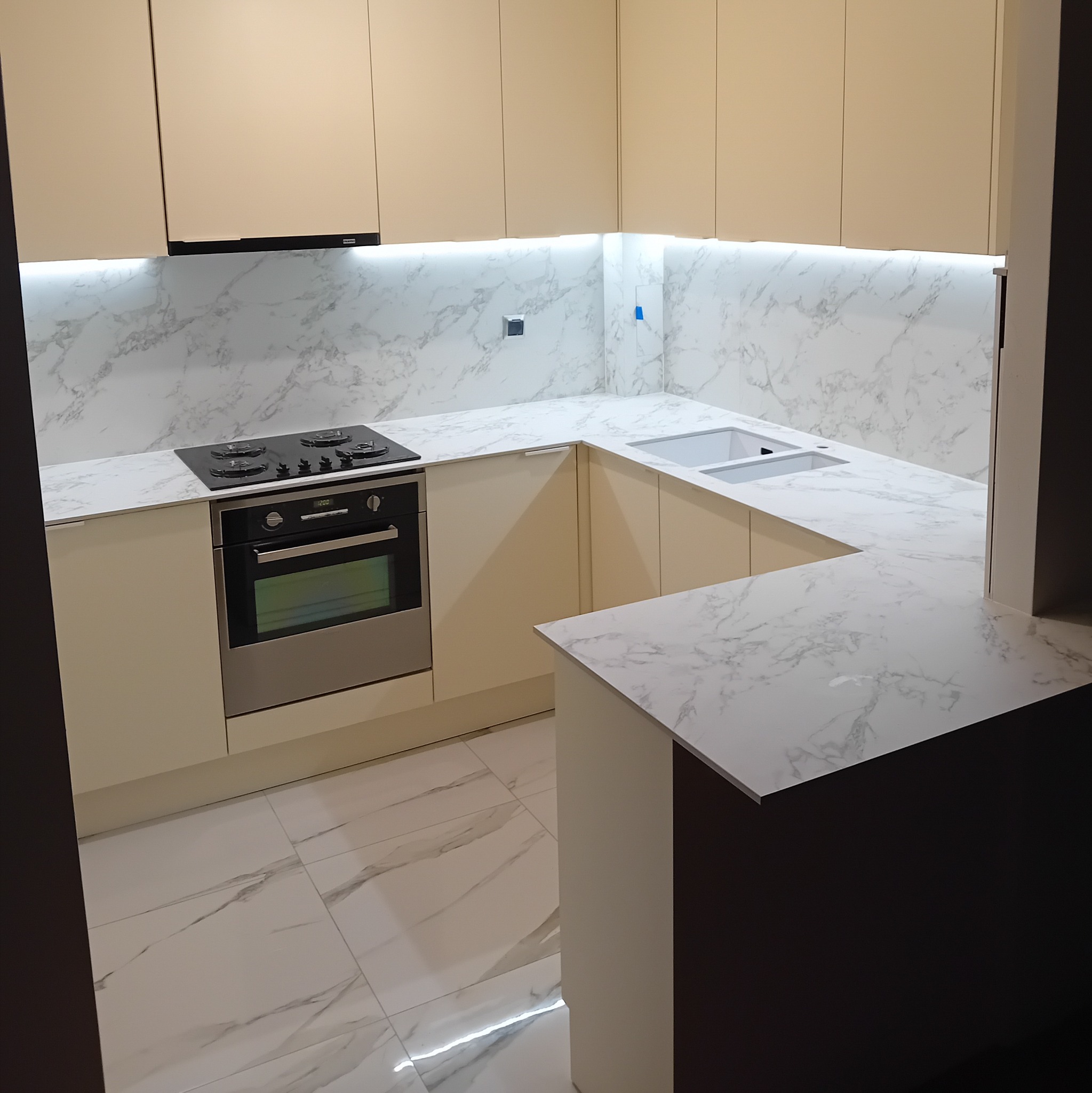Nowoczesna kuchnia w kształcie litery U z beżowymi szafkami, marmurowym blatem i podświetleniem LED pod szafkami wiszącymi, widoczna płyta grzewcza i piekarnik ze stali nierdzewnej.