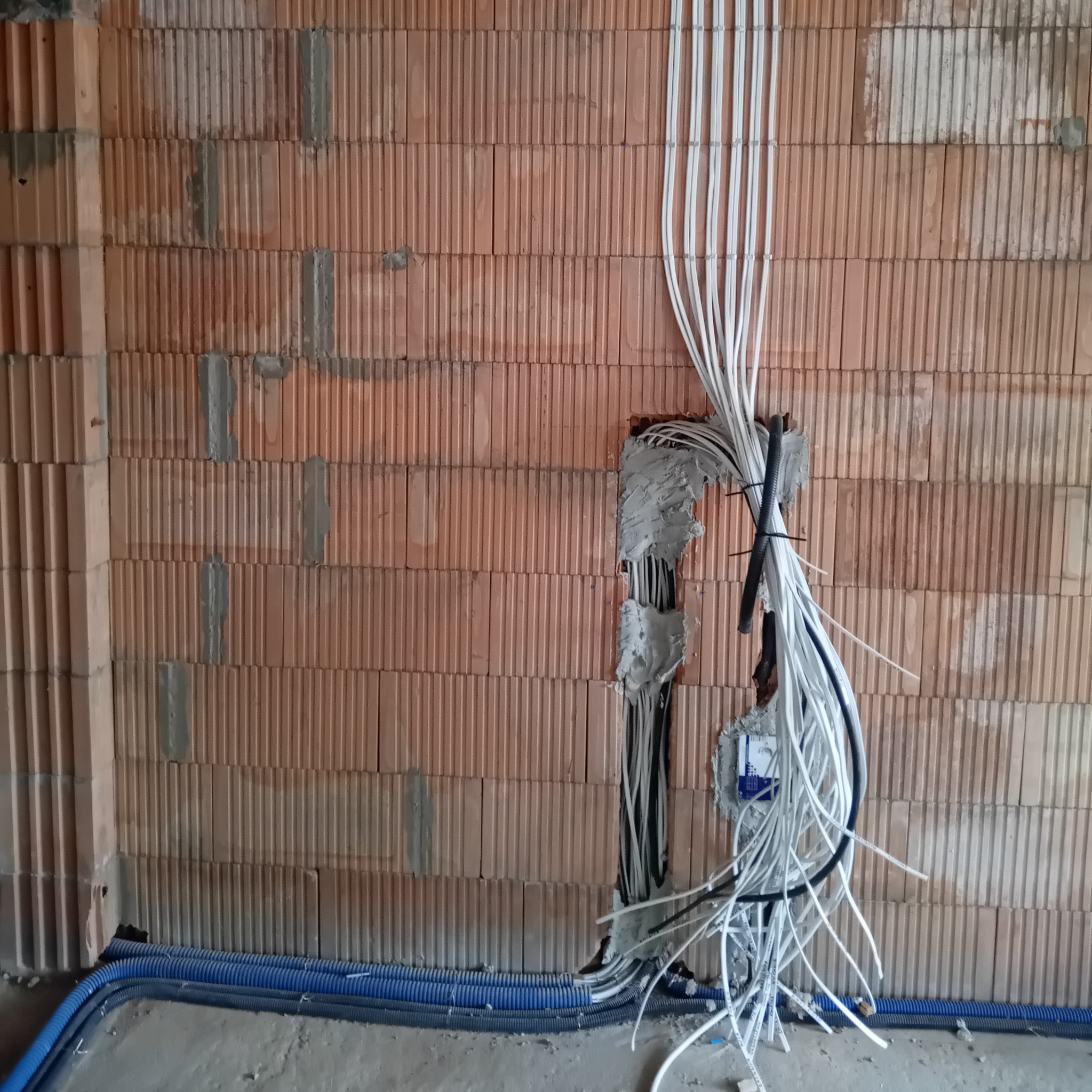 Wiązka przewodów elektrycznych w białej izolacji, wychodząca ze ściany z czerwonej cegły dziurawki, zabezpieczona gipsem, z widocznymi niebieskimi rurami karbowanymi w dolnej części kadru.