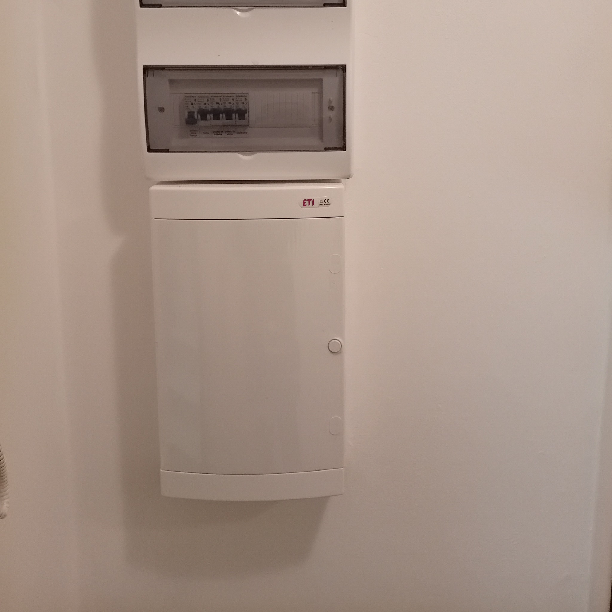 Zamknięta, biała skrzynka elektryczna ETI z widocznymi bezpiecznikami na białej ścianie.