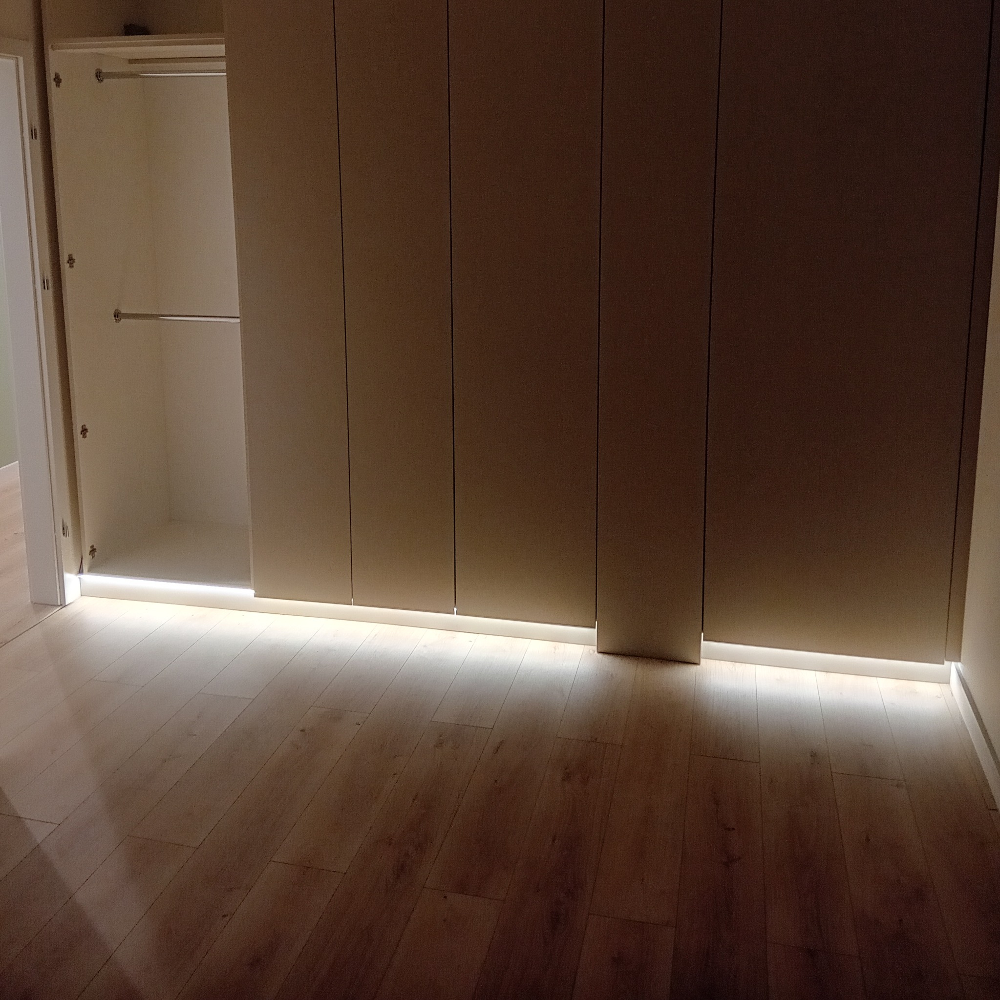 Wnętrze szafy wnękowej z oświetleniem LED u dołu, widok na drewnianą podłogę i część otwartej szafy z drążkami na ubrania.