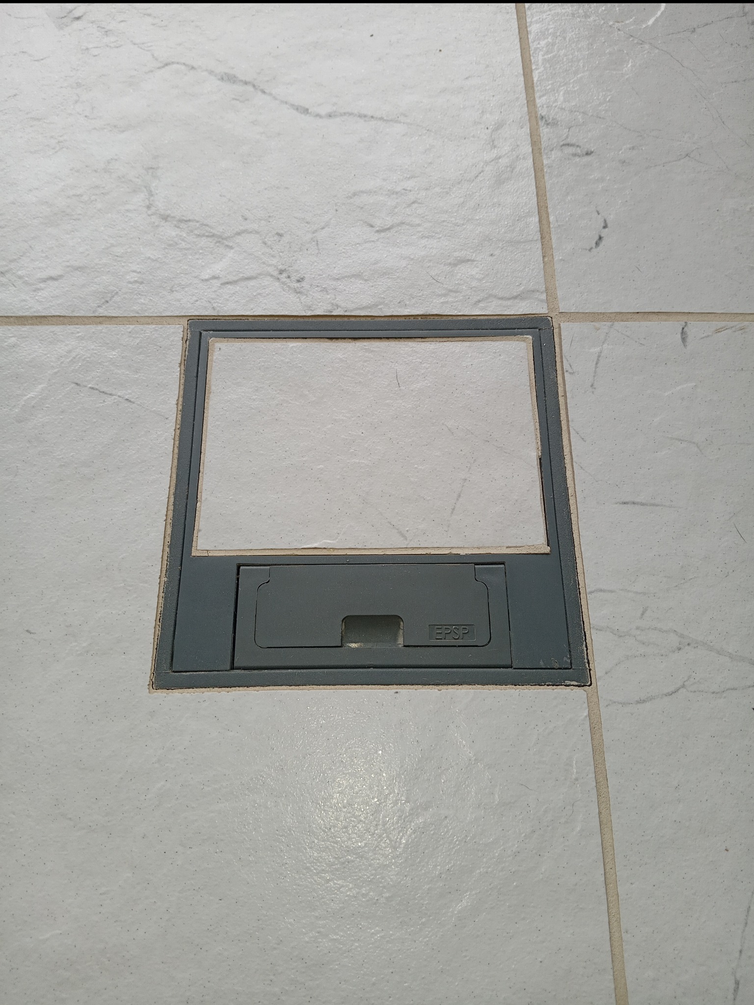 Szara podłogowa puszka elektryczna EPSP wkomponowana w jasną, marmurkową podłogę z płytek, widok z góry.