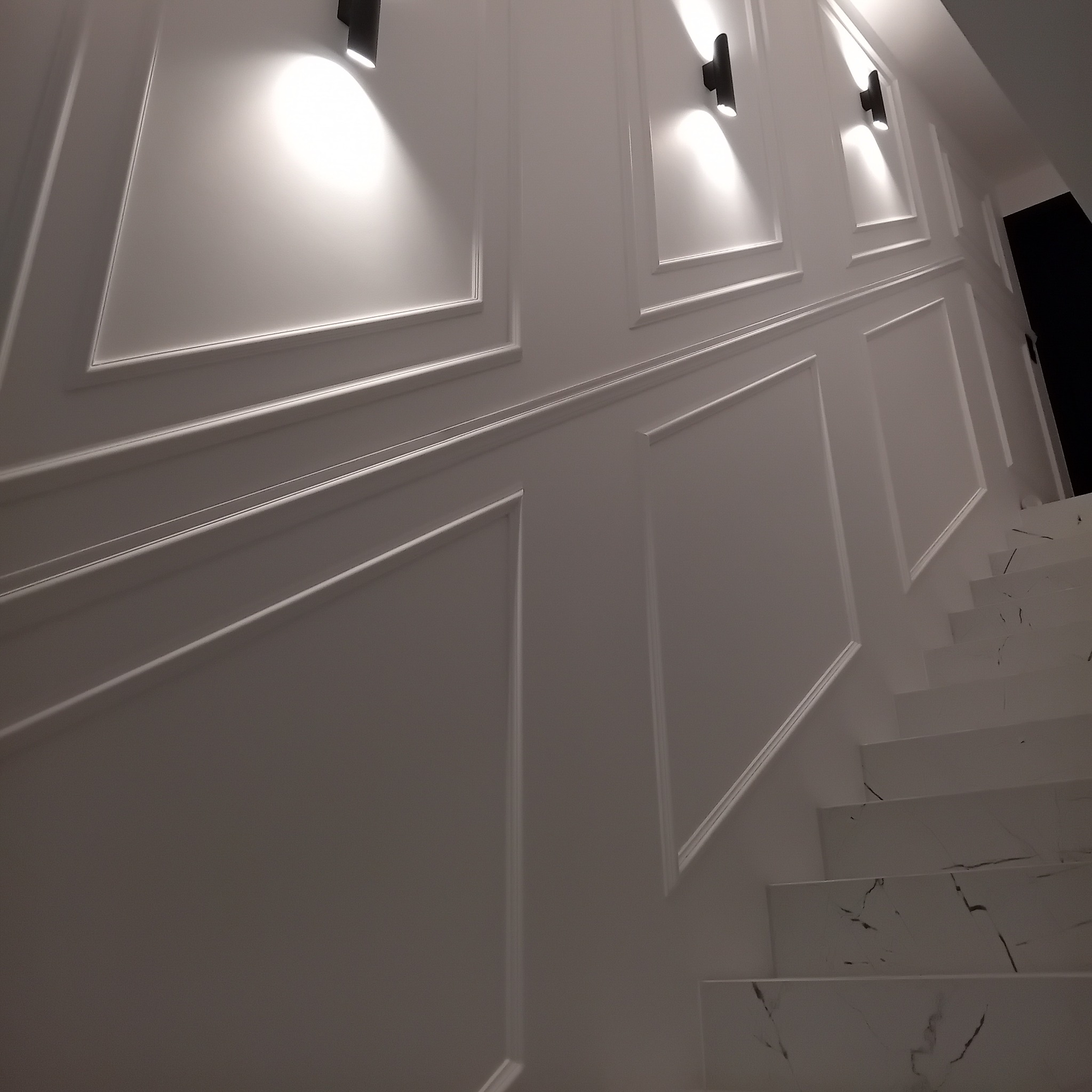 Nowoczesne oświetlenie schodów z kinkietami LED na białej ścianie z dekoracyjnymi panelami.