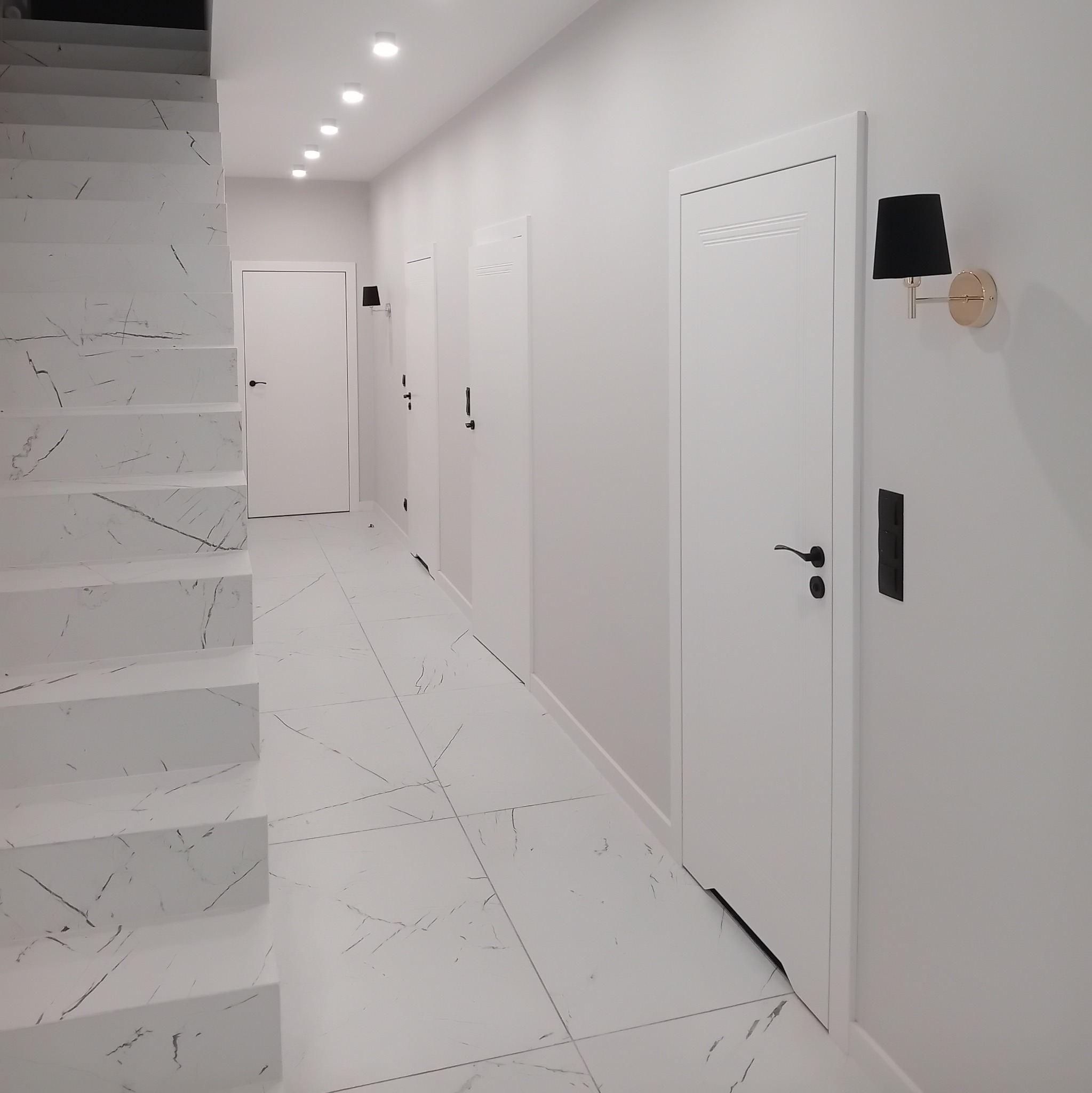 Nowoczesny korytarz w jasnych barwach z marmurowymi schodami, białymi drzwiami z czarnymi klamkami i kinkietami z czarnymi abażurami.