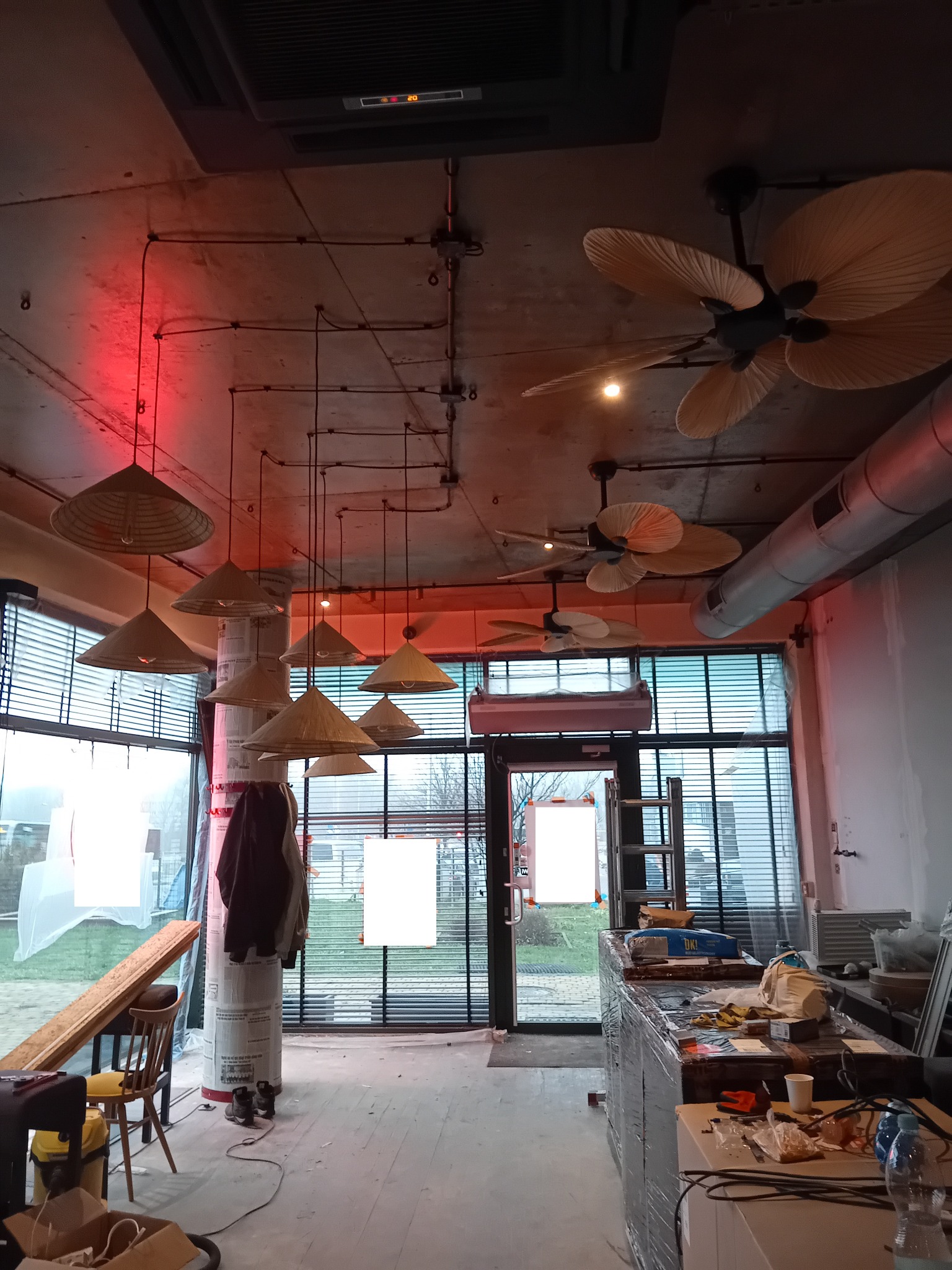 Wnętrze lokalu w trakcie wykańczania, widoczne lampy w kształcie kapeluszy, wentylatory sufitowe, odkryte przewody elektryczne pod sufitem, elementy instalacji wentylacyjnej, okna z żaluzjami...