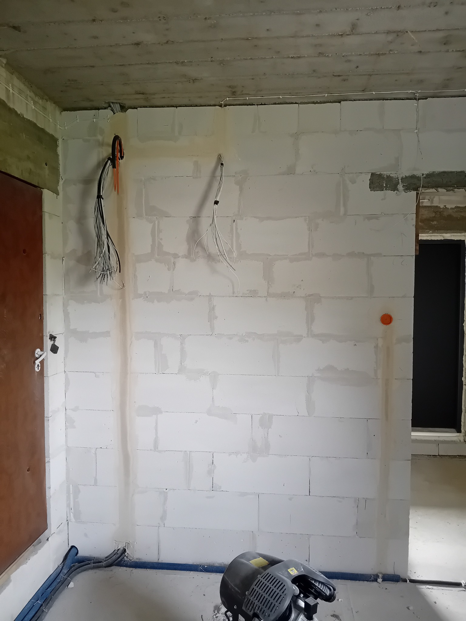 Wnętrze w trakcie instalacji elektrycznej: przewody zwisają ze ściany z białego bloczka, niebieskie rury w podłodze, widoczny agregat. Etap prac wykończeniowych.