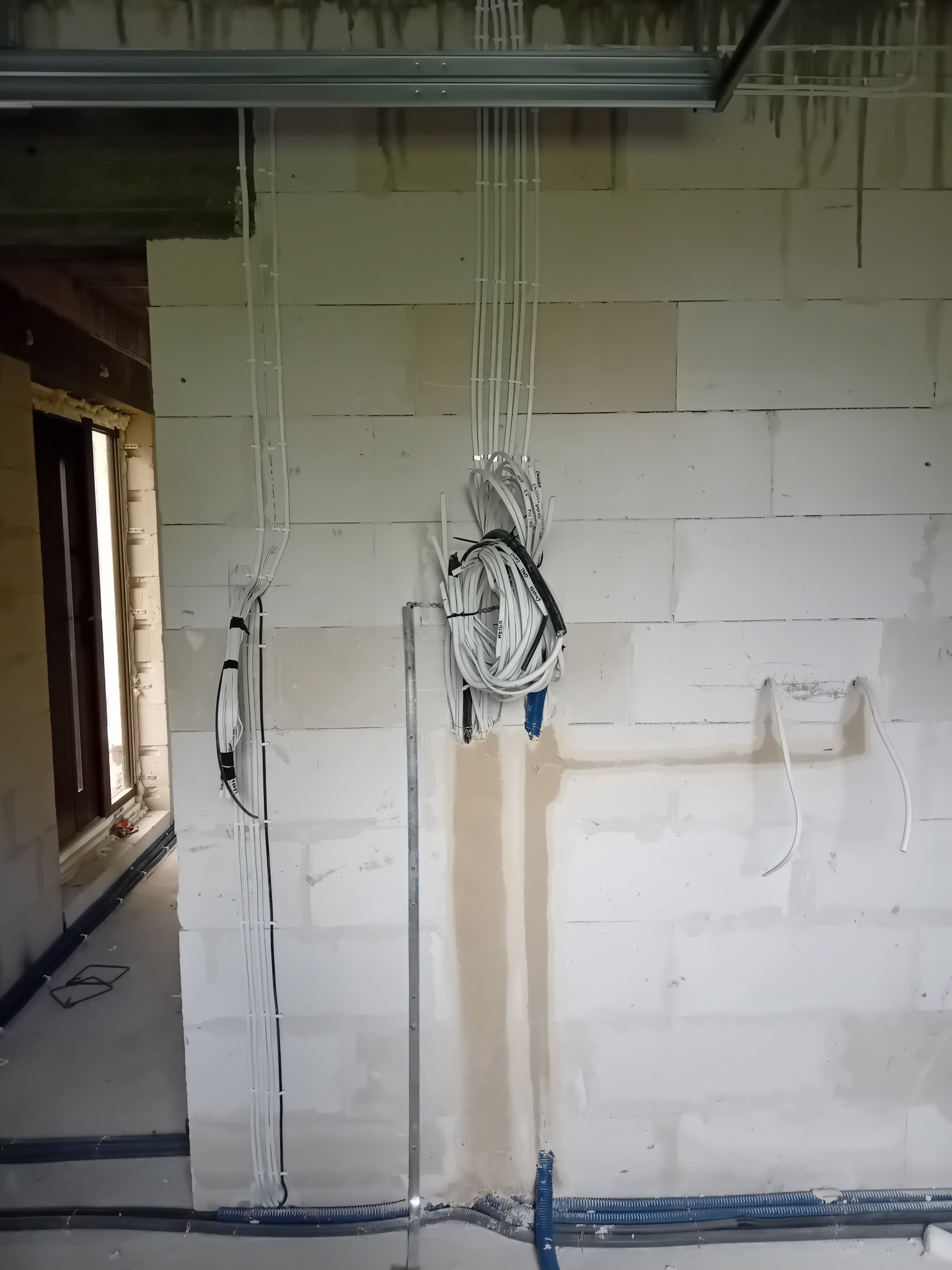 Surowe wnętrze z instalacją elektryczną: białe kable prowadzone po ścianach z betonu komórkowego, częściowo w peszlach pod podłogą. Widoczne elementy konstrukcji sufitu.