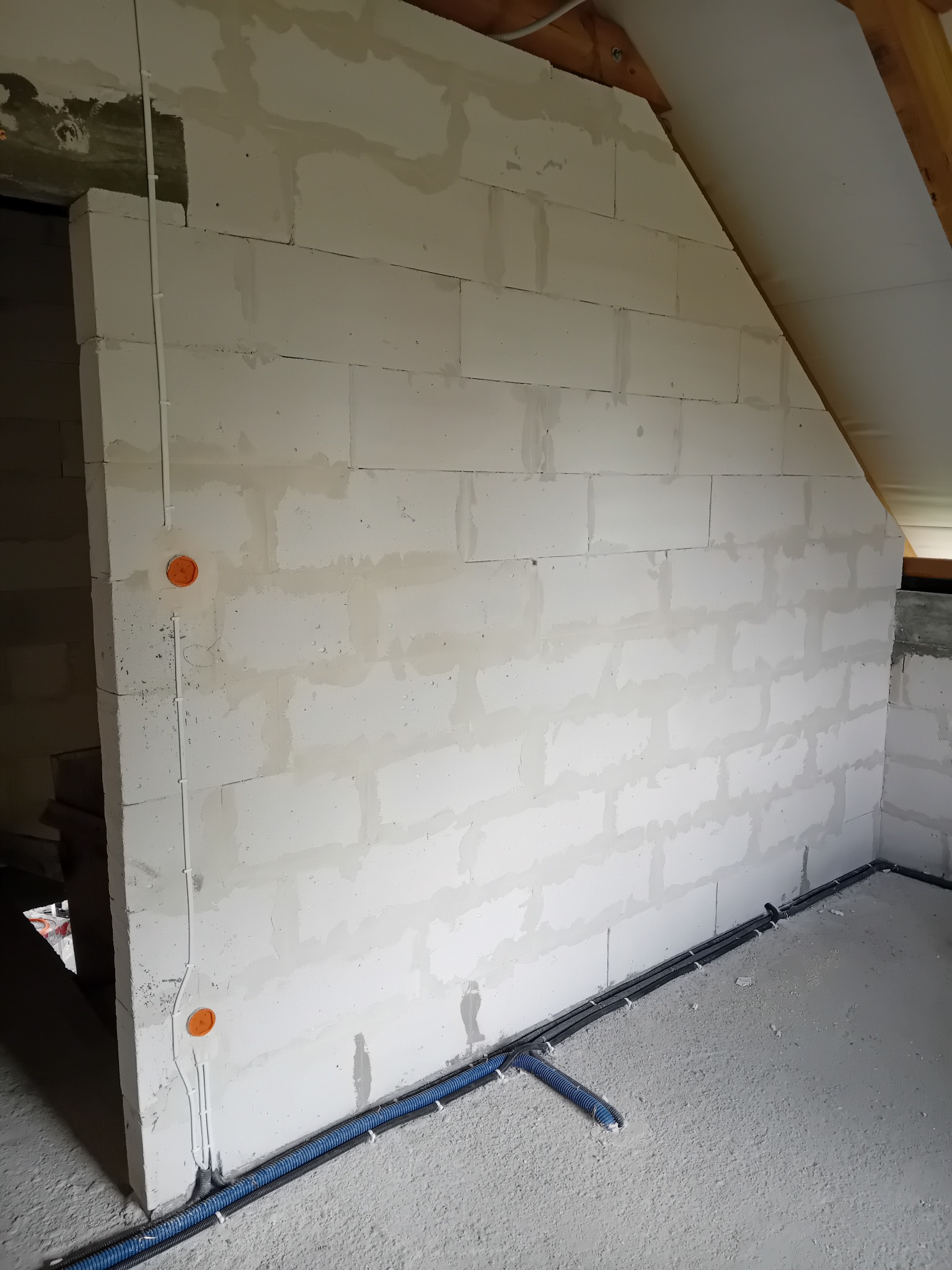 Surowy stan poddasza z instalacją elektryczną w peszlach przy ścianie z betonu komórkowego, widoczne puszki instalacyjne i przewody. Etap prac instalacyjnych.