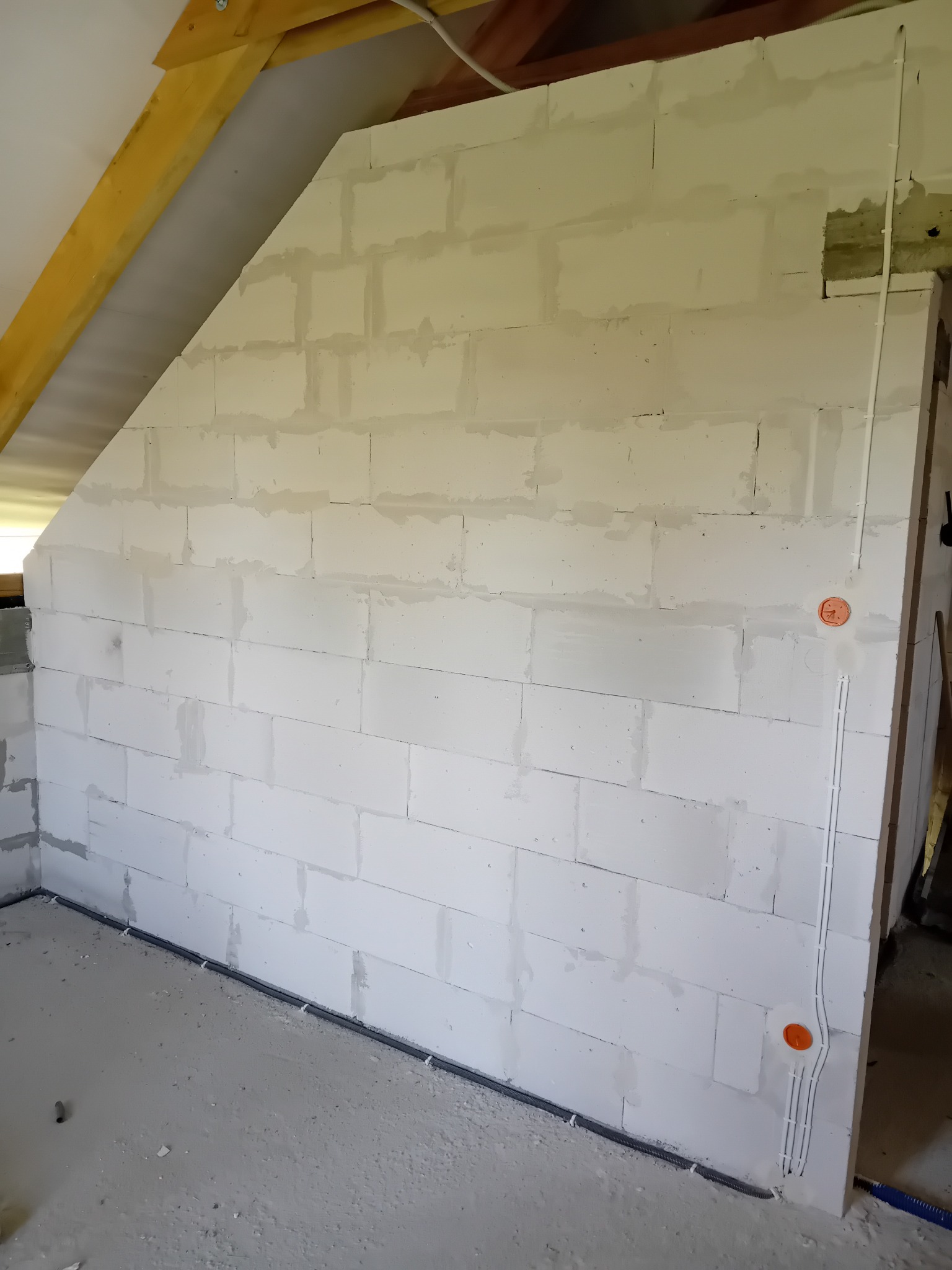 Surowa ściana z betonu komórkowego na poddaszu z widoczną instalacją elektryczną w peszlach i puszkami. Widoczne elementy konstrukcji dachu.