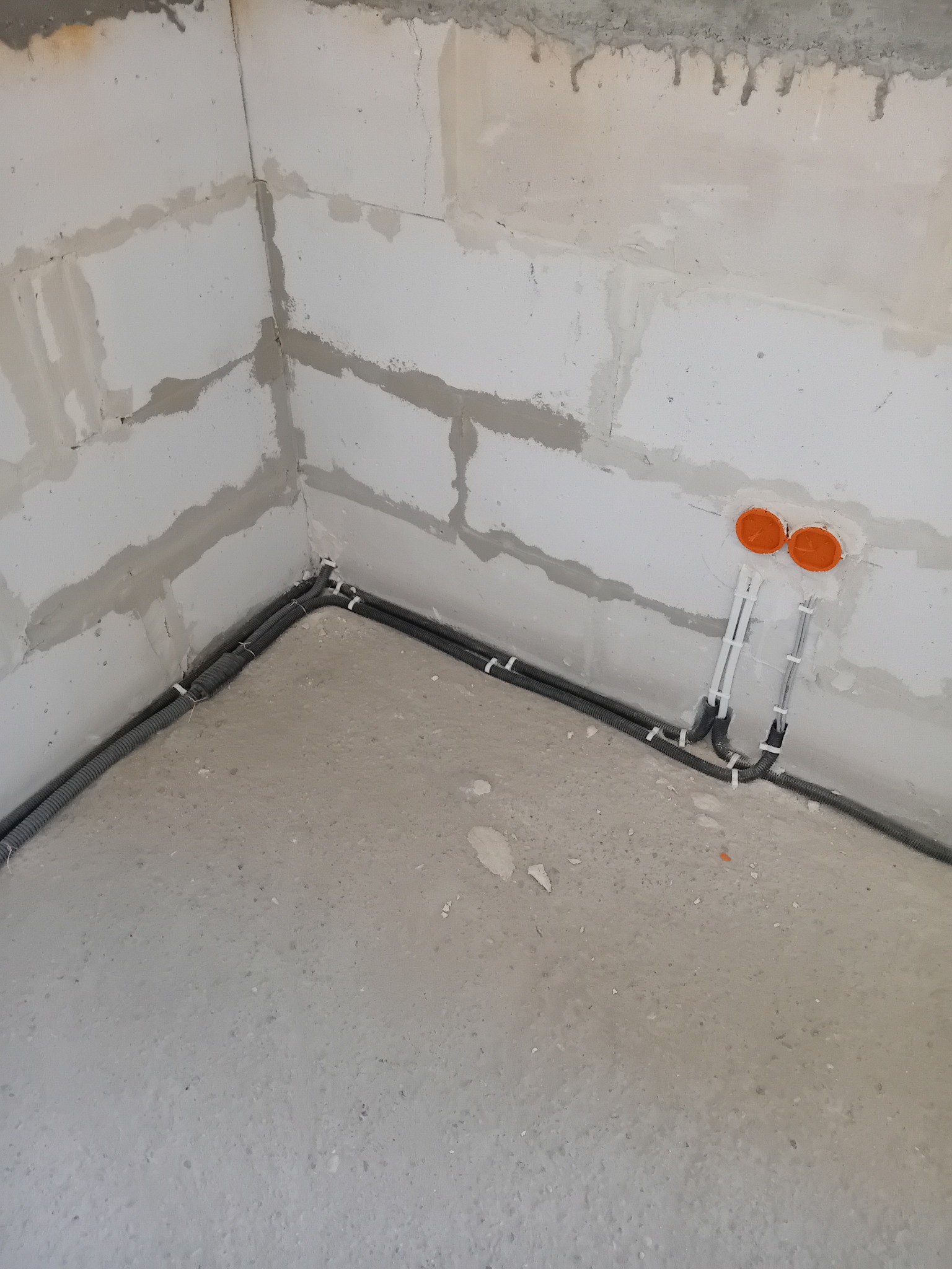 Szare rury w peszlu poprowadzone wzdłuż ściany z bloków. Widoczne pomarańczowe puszki instalacyjne. Instalacja elektryczna w surowym stanie.