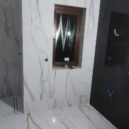 Wnętrze łazienki w trakcie prac wykończeniowych, z białymi płytkami imitującymi marmur z złotymi żyłami na ścianach i podłodze, ciemne płytki w strefie prysznica, drewniane okno z widocznymi...