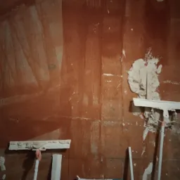 Czerwona ściana w trakcie przygotowania do dalszych prac wykończeniowych, widoczne ślady po szpachlowaniu i narzędzia, worek z materiałem budowlanym w dolnej części kadru.