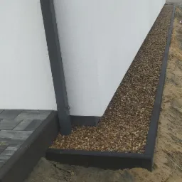 Obrzeże z kostki brukowej i kamienny drenaż przy ścianie budynku z rynną spustową, widok z poziomu gruntu.