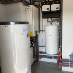 Nowa instalacja z zbiornikiem ciepłej wody owiniętym folią ochronną, obok pompa ciepła i elementy instalacji grzewczej z rurami w izolacji, widoczne etykiety energetyczne i napisy na rurach...