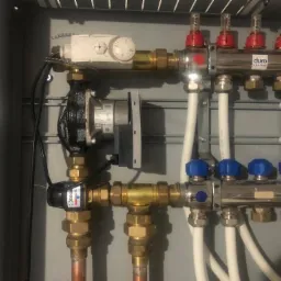 Rozdzielacz podłogowy z pompą obiegową Wilo, zaworami termostatycznymi z czerwonymi pokrętłami i niebieskimi zaworami kulowymi, zamontowany w szarej szafce. Widoczne miedziane rury i białe rurki...